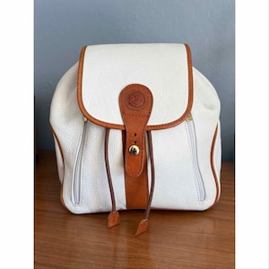 May include: A white backpack with a pebbled texture and tan leather trim. The bag features a fold-over flap with a central tan leather strap, a gold-tone metal snap closure, and two thin drawstring cords. Two vertical zippered pockets are on the front.