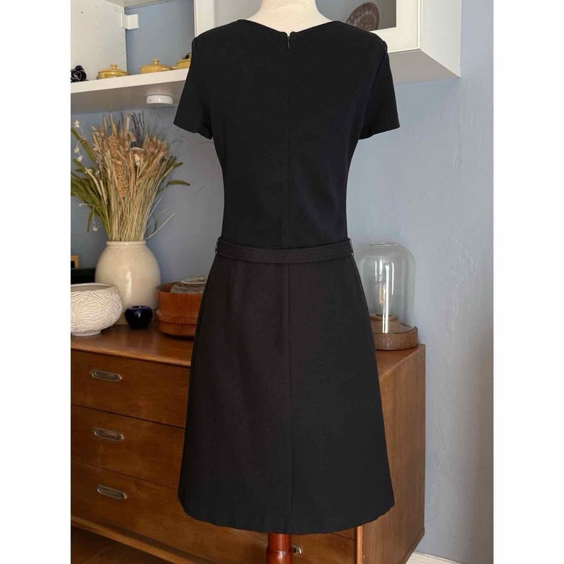 May include: A black dress with short sleeves and a fitted waist. The dress has a simple, classic design, with a subtle belt detail at the waist. The dress is displayed on a mannequin, showcasing its shape and style. The dress is a versatile piece suitable for various occasions.
