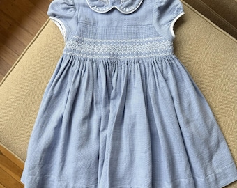 Puede incluir: Vestido de bebé azul claro con cuello blanco y mangas cortas. El vestido presenta un diseño bordado blanco detallado en el pecho y una falda fruncida. La tela tiene un sutil patrón texturizado.