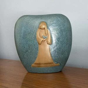 Puede incluir: Un jarrón de cerámica con forma de corazón con un diseño en relieve de una mujer sosteniendo un pájaro. El jarrón es de color azul claro con un esmalte marrón en la mujer y el pájaro.