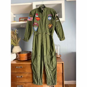 May include: Olive green military jumpsuit with various patches. The jumpsuit features a zip closure, long sleeves, and pockets on the chest and legs. Patches include an American flag, a red and yellow patch, and a circular patch with a gold emblem.