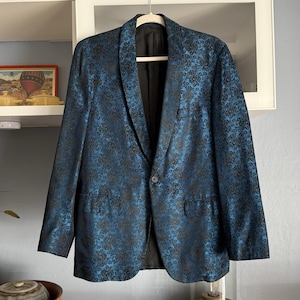Peut inclure: Un blazer bleu avec un motif floral noir. La veste a une fermeture à un bouton, un revers cranté et deux poches avant. Le blazer est suspendu à un cintre en bois.