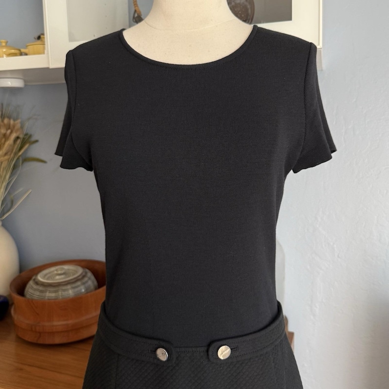 May include: A black dress with a ribbed bodice and short sleeves. The skirt features a quilted pattern and is accented with two silver buttons. The dress is displayed on a mannequin.