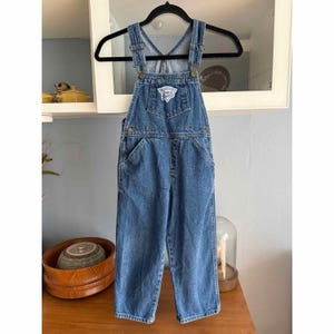 May include: Blue denim overalls with adjustable straps and pockets. The overalls have a front bib pocket with a small patch. The garment is hanging on a black hanger.