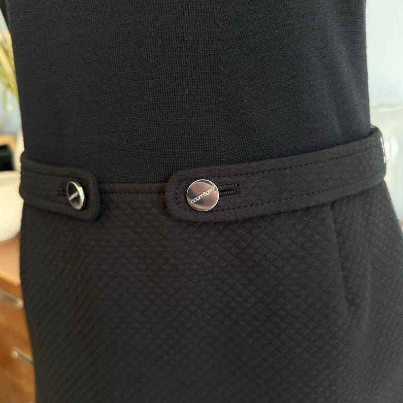 May include: Black dress with a textured skirt and a fitted waist. The dress features a matching belt with a silver-tone button closure. The button is embossed with the word "courr&egrave;ges".
