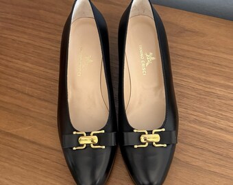May include: A pair of black leather flats with pointed toes. Each shoe features a gold-colored buckle detail on the vamp. The interior of the shoes is a light beige color. The shoes are displayed on a wooden surface.