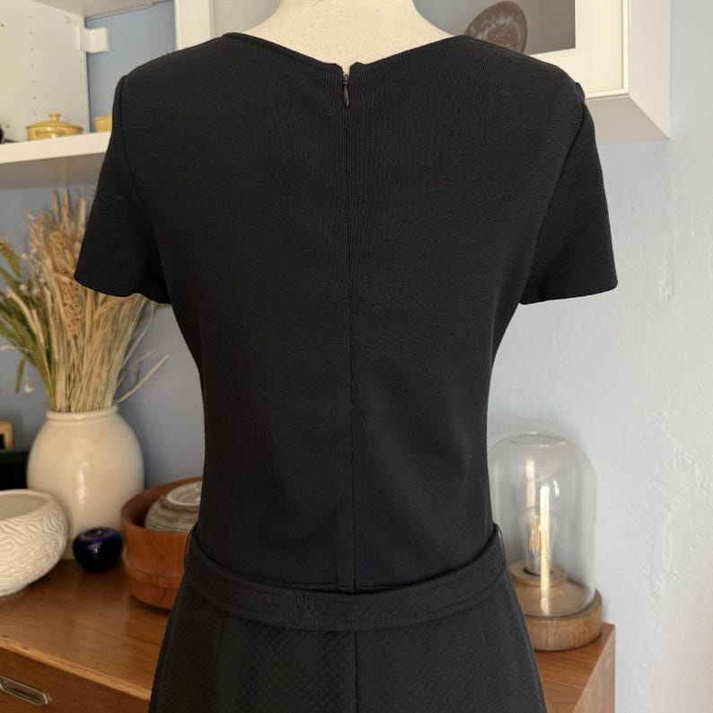 May include: A black dress with short sleeves and a textured pattern. The dress features a fitted waist and a zipper closure at the back. The dress is displayed on a mannequin.