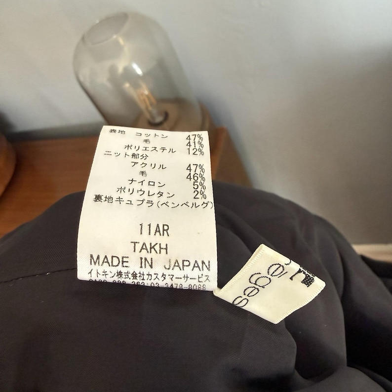 May include: Close-up of a black garment tag with Japanese text and fabric composition percentages. The tag reads "11AR TAKH MADE IN JAPAN". A small white tag with the brand name "sabe" is also visible.