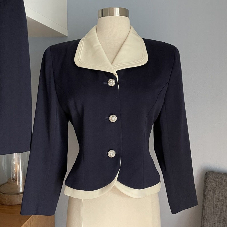 K&ouml;nnte beinhalten: Ein marineblauer Blazer mit wei&szlig;em Kragen und wei&szlig;em Besatz an den R&auml;ndern. Der Blazer hat vier wei&szlig;e Kn&ouml;pfe mit Strasssteinen in der Mitte.