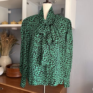 May include: A green blouse with a black oval pattern. The long-sleeved top features a high collar and a large bow at the neckline. The blouse is displayed on a mannequin.