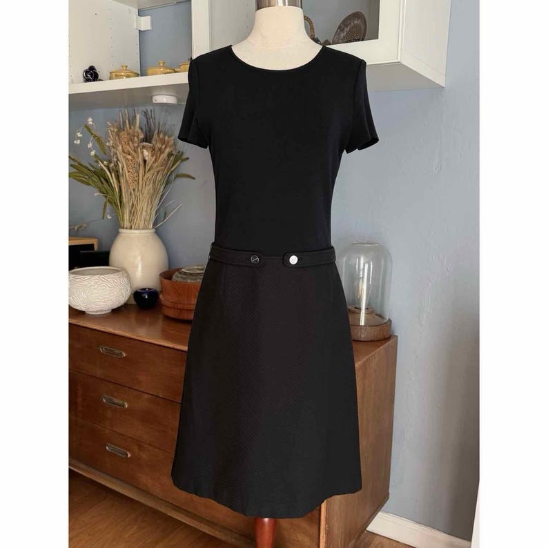May include: A black dress with short sleeves and a fitted waist. The dress features a round neckline and a skirt that falls to the knees. The waist is accented with a thin belt and a single button closure. The dress is displayed on a mannequin.