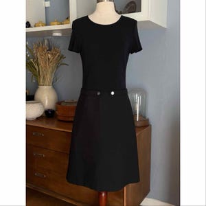 May include: A black dress with short sleeves and a round neckline. The dress features a fitted waist with two decorative buttons. The skirt is a-line, and the dress is displayed on a mannequin.