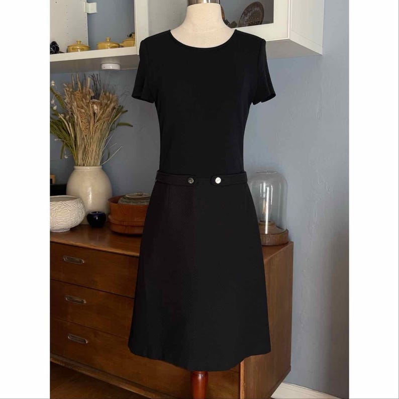 May include: A black dress with short sleeves and a round neckline. The dress features a fitted waist with two decorative buttons. The skirt is a-line, and the dress is displayed on a mannequin.