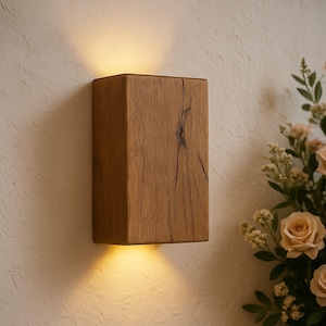 May include: A rectangular wooden wall sconce with a warm, natural wood grain finish. The sconce emits a soft, downward-facing light, creating a cozy ambiance. The design features a prominent crack in the wood, adding character. The sconce is mounted on a textured, off-white wall.