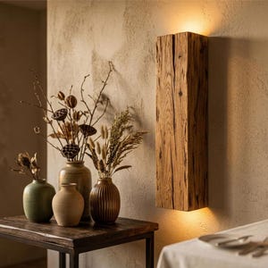 May include: A wooden wall light fixture, emitting a warm glow. The light is made from a rectangular block of weathered wood, mounted on a beige wall. Decorative vases with dried flowers sit on a dark wooden table.