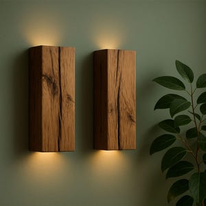 May include: Two rectangular wooden wall sconces with a warm, glowing light emanating from the bottom. The sconces are made of textured, light brown wood and mounted on a muted green wall. A leafy green plant is visible on the right side.