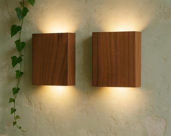 以下が含まれることがあります： 下部から温かい光を発する、長方形の木製ウォールスコン2つ。 スコンは天然の木目仕上げで、質感のある淡い緑色の壁に取り付けられています。 緑の葉を持つつる植物が見えます。