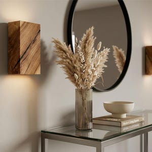 May include: Two wooden wall sconces with a warm glow, flanking a decorative arrangement. The sconces are rectangular with a natural wood grain pattern. A glass-topped table holds a vase of dried flowers, a bowl, and a book.