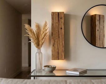 May include: A rustic wooden wall sconce with a warm, glowing light. The sconce is made of a thick, textured piece of wood. A round black mirror and a glass table with decor complete the scene. A vase of dried flowers and a small bowl are also visible.