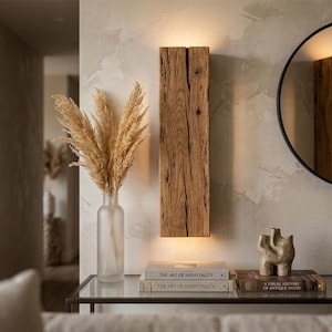 May include: A rustic wooden wall sconce illuminates a neutral-toned room. A frosted glass vase holds dried pampas grass. A black-framed mirror and decorative objects sit on a glass-topped table. Books with the title "The Art of Hospitality" are visible.