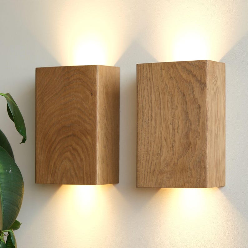 Op de afbeelding: Twee houten wandlampen met een warm licht dat van onderaan elke lamp straalt. De lampen zijn gemaakt van natuurlijk hout met een lichtbruine afwerking.