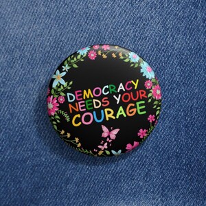 May include: A round black button with the text "DEMOCRACY NEEDS YOUR COURAGE" in rainbow colors, surrounded by a floral wreath. The button is set against a blue denim jacket.