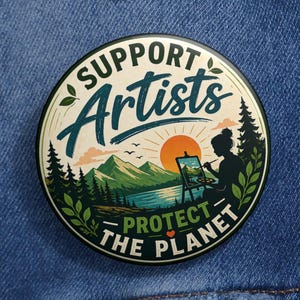 May include: A round button with the text "Support Artists Protect the Planet." The design features a landscape with mountains, trees, a lake, and a setting sun. A silhouette of a person painting is also included.