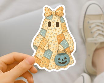 Peut inclure: Un autocollant représentant un fantôme avec un motif de patchwork jaune, orange et bleu. Le fantôme porte un nœud et un seau citrouille bleu. L'autocollant est tenu par une main, avec un ordinateur portable et des baskets en arrière-plan.