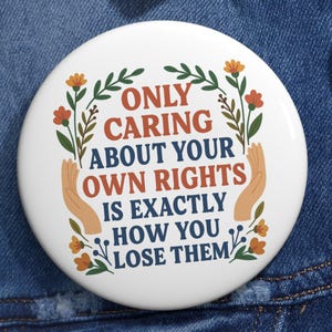 Può includere: Un bottone bianco con la scritta "ONLY CARING ABOUT YOUR OWN RIGHTS IS EXACTLY HOW YOU LOSE THEM" in marrone e arancione. Il bottone è decorato con illustrazioni floreali e di mani, su una giacca di jeans.