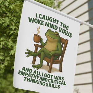 May include: White flag with a green frog illustration, seated in a wooden chair and holding a mug. The flag has the text "I CAUGHT THE WOKE MIND VIRUS AND ALL I GOT WAS EMPATHY AND CRITICAL THINKING SKILLS" in green font.