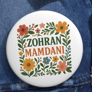 May include: A white button with the name "Zohran Mamdani" in green and orange text, surrounded by colorful flowers and green leaves. The button is set against a blue denim jacket.