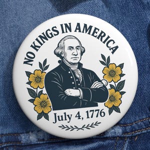 Puede incluir: Un botón blanco con el texto "NO KINGS IN AMERICA" que rodea un retrato de George Washington. El botón también presenta la fecha "July 4, 1776" y detalles florales amarillos, sobre una chaqueta vaquera.
