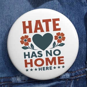 May include: A white button with the words "HATE HAS NO HOME HERE" in a bold, stylized font. The design includes a teal heart, orange flowers, and stars. The button is pinned to a blue denim jacket.