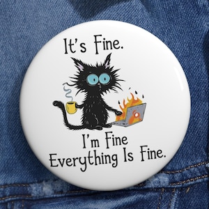 Puede incluir: Un botón blanco con una ilustración de un gato negro sosteniendo una taza y usando una computadora portátil en llamas. El texto dice: "It's Fine. I'm Fine. Everything Is Fine."