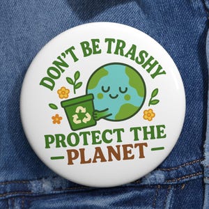 May include: A white button with the text "DON'T BE TRASHY" and "PROTECT THE -PLANET-". The button features a smiling Earth holding a recycling bin, with green leaves and orange flowers. The button is on a denim jacket.