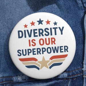 May include: A white badge with the words "DIVERSITY IS OUR SUPERPOWER" in navy and red. The design includes stars and a wing motif in red, navy, and tan, set against a denim jacket.