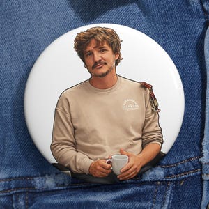 Op de afbeelding: Een witte button met een portret van een man in een beige sweatshirt die een mok vasthoudt. Het sweatshirt heeft een cirkelvormig logo met tekst. De button is geplaatst op een blauw spijkerjasje.