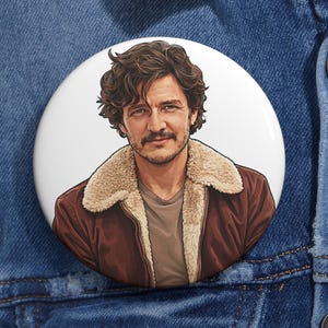 Op de afbeelding: Een witte button met een portret van een man met bruin haar en een snor, gekleed in een bruine jas met een beige bontkraag. De button is geplaatst tegen een blauwe denim achtergrond.