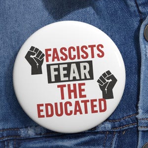 May include: White badge with the text "Fascists Fear The Educated" in black and red, featuring black fist graphics. The badge is pinned to a blue denim jacket.