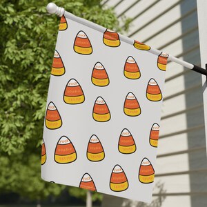 May include: A white flag with a repeating pattern of candy corn with the text "FUCK DONALD TRUMP" in orange and yellow. The flag is mounted on a white wall with a black bracket.