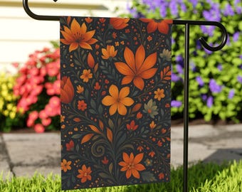 May include: A decorative garden flag featuring a floral pattern in shades of orange, rust, and green on a dark background. The flag is displayed on a black metal stand, adding a touch of seasonal charm to outdoor spaces.