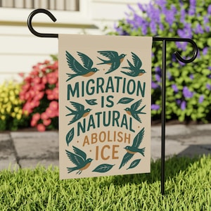 May include: A beige garden flag with the text "MIGRATION IS NATURAL ABOLISH ICE" in teal and orange. The flag features teal birds and leaves. The flag is displayed on a black metal stand in a garden setting.