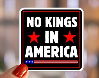 May include: A square sticker with the text "NO KINGS IN AMERICA" in white, set against a black background with red stars and a red and white striped design. The sticker is held by a hand with red nail polish.