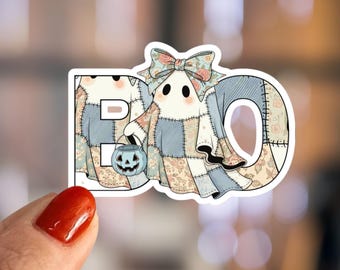 Peut inclure: Autocollant avec le mot "BO" représentant un fantôme et un seau citrouille. Le fantôme porte un costume en patchwork avec un nœud floral. L'autocollant a une bordure blanche et est tenu par une main.