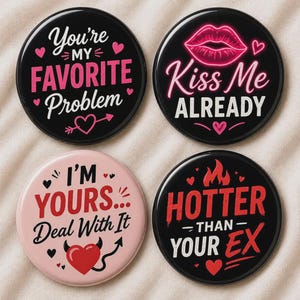 Könnte beinhalten: Vier runde schwarze und rosa Knöpfe mit romantischen Phrasen. Die Knöpfe zeigen Sätze wie "You're My Favorite Problem", "Kiss Me Already", "I'm Yours... Deal With It" und "Hotter Than Your Ex."