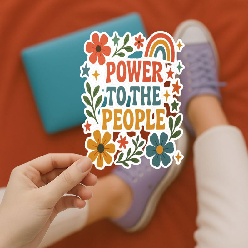 K&ouml;nnte beinhalten: Ein farbenfroher Aufkleber mit dem Text "POWER TO THE PEOPLE", umgeben von Blumen- und Regenbogenmotiven. Der Aufkleber wird von einer Hand gehalten, mit einem blauen Laptop und lila Schuhen im Hintergrund. Der Aufkleber hat orange, gelbe, gr&uuml;ne und blaue Farben.