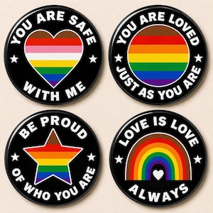 Puede incluir: Conjunto de cuatro botones redondos negros con texto blanco y gráficos de arcoíris. Los botones presentan frases como "Eres amado tal como eres" y "El amor es amor siempre".