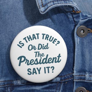 May include: A white badge with the teal text "IS THAT TRUE? Or Did The President SAY IT?". The badge is pinned to a blue denim jacket.