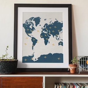 May include: A framed print of a world map with a blue and white color scheme. The map is decorated with small gold airplane icons.