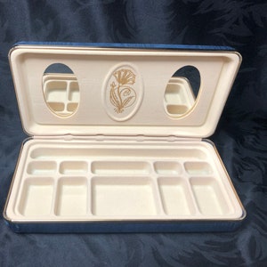 Puede incluir: Una caja de joyería de terciopelo azul con ribete dorado y dos espejos. La caja tiene un diseño floral en la tapa y un compartimento con múltiples secciones para guardar joyas.